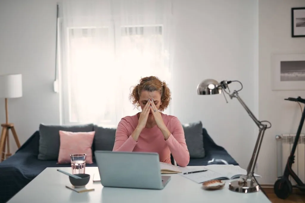 Person with reddish-brown curly hair wearing a pink top, sitting at a desk with hands to face in a stressed pose, with laptop, papers, and a glass of water visible