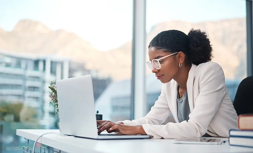 Woman wearing glasses and beige blazer typing on laptop at desk with mountain view through window