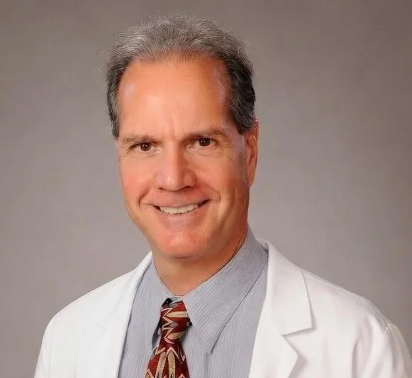 Middle-aged man with gray hair wearing white lab coat, light blue striped shirt and burgundy tie, smiling at camera against neutral gray background.