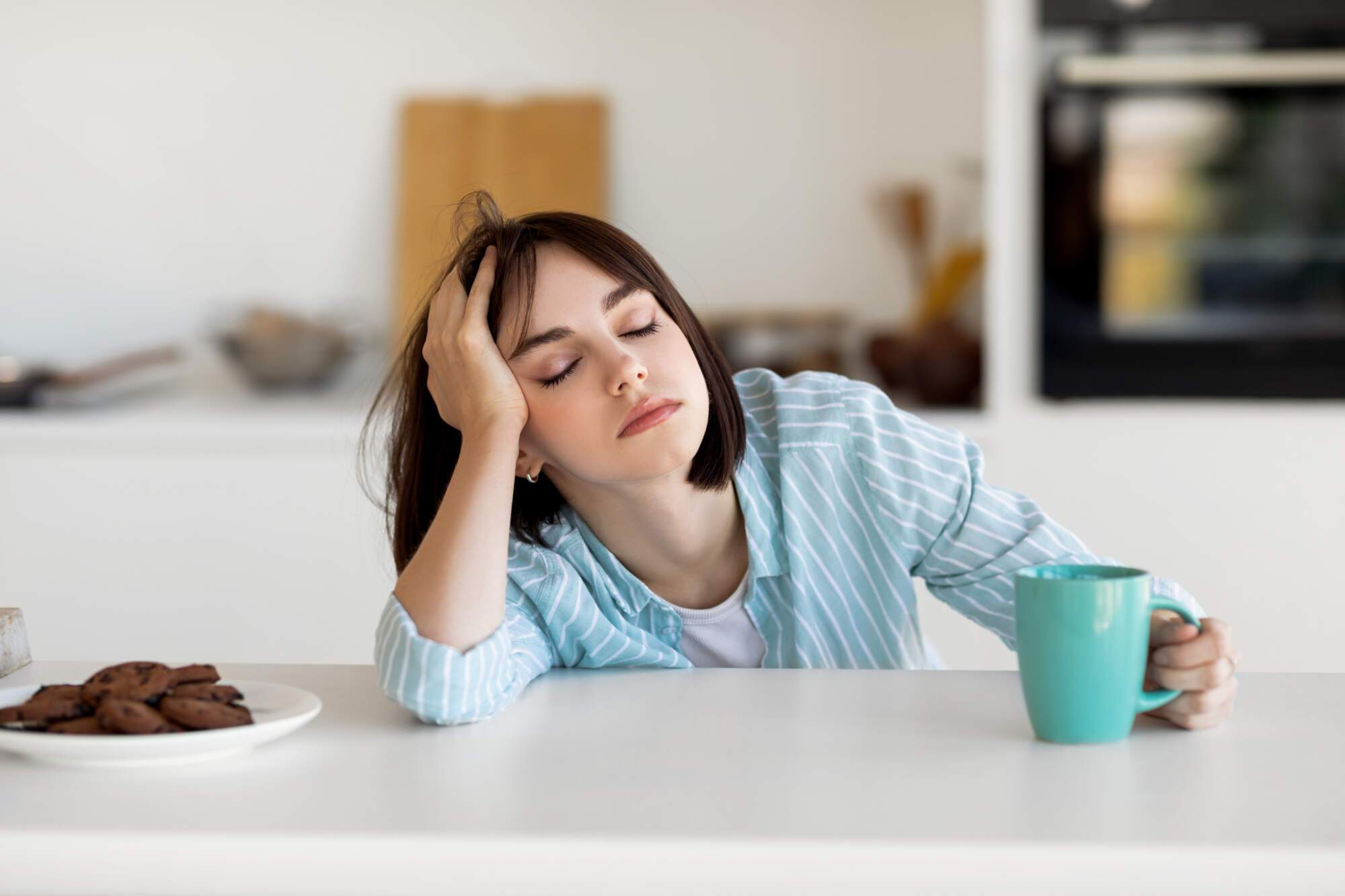 Woman with dark hair resting her head on her hand at a kitchen table, looking tired and holding a turquoise mug, with a plate of cookies nearby