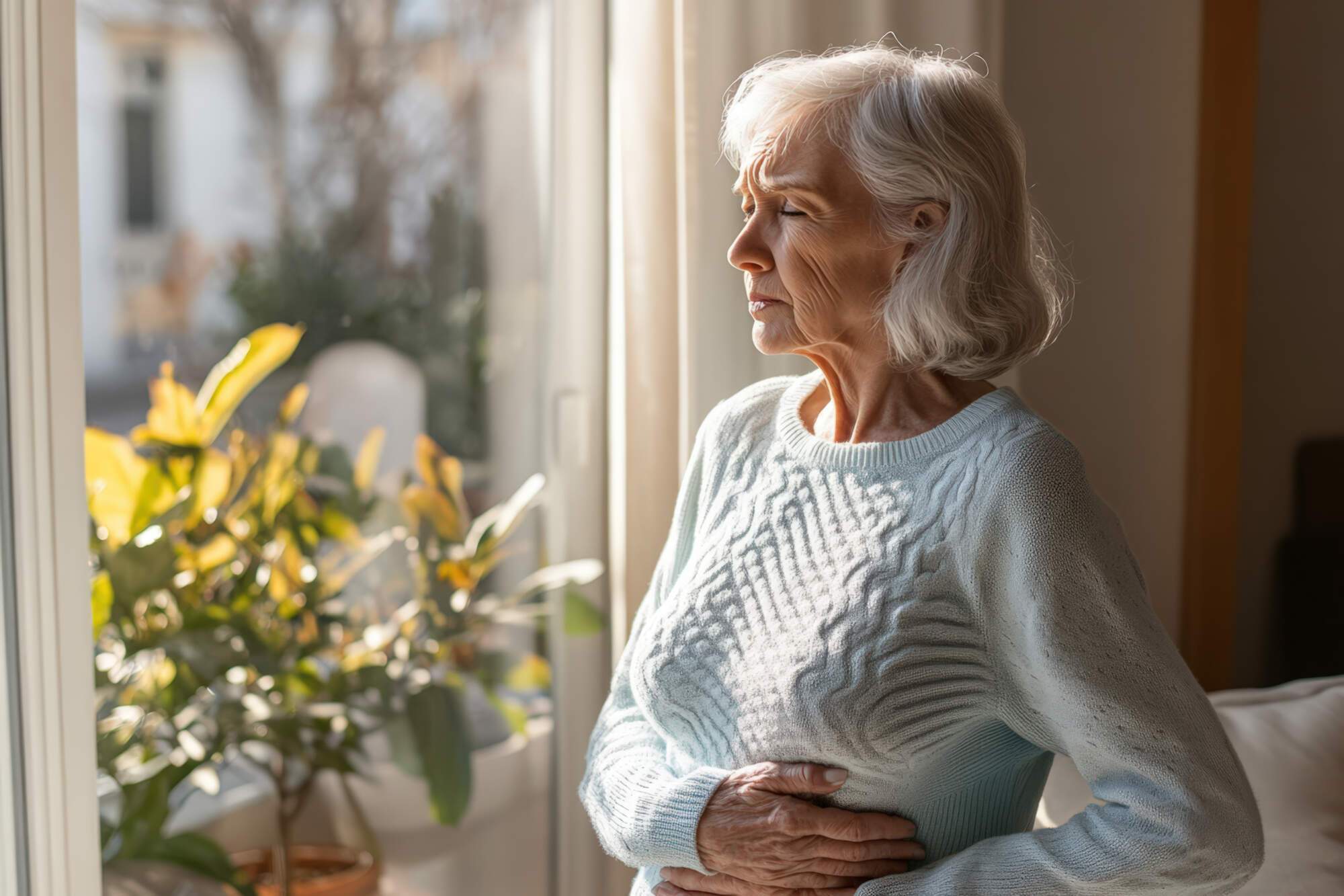 Older woman with gray hair wearing a light blue sweater looking out a window with a potted plant nearby