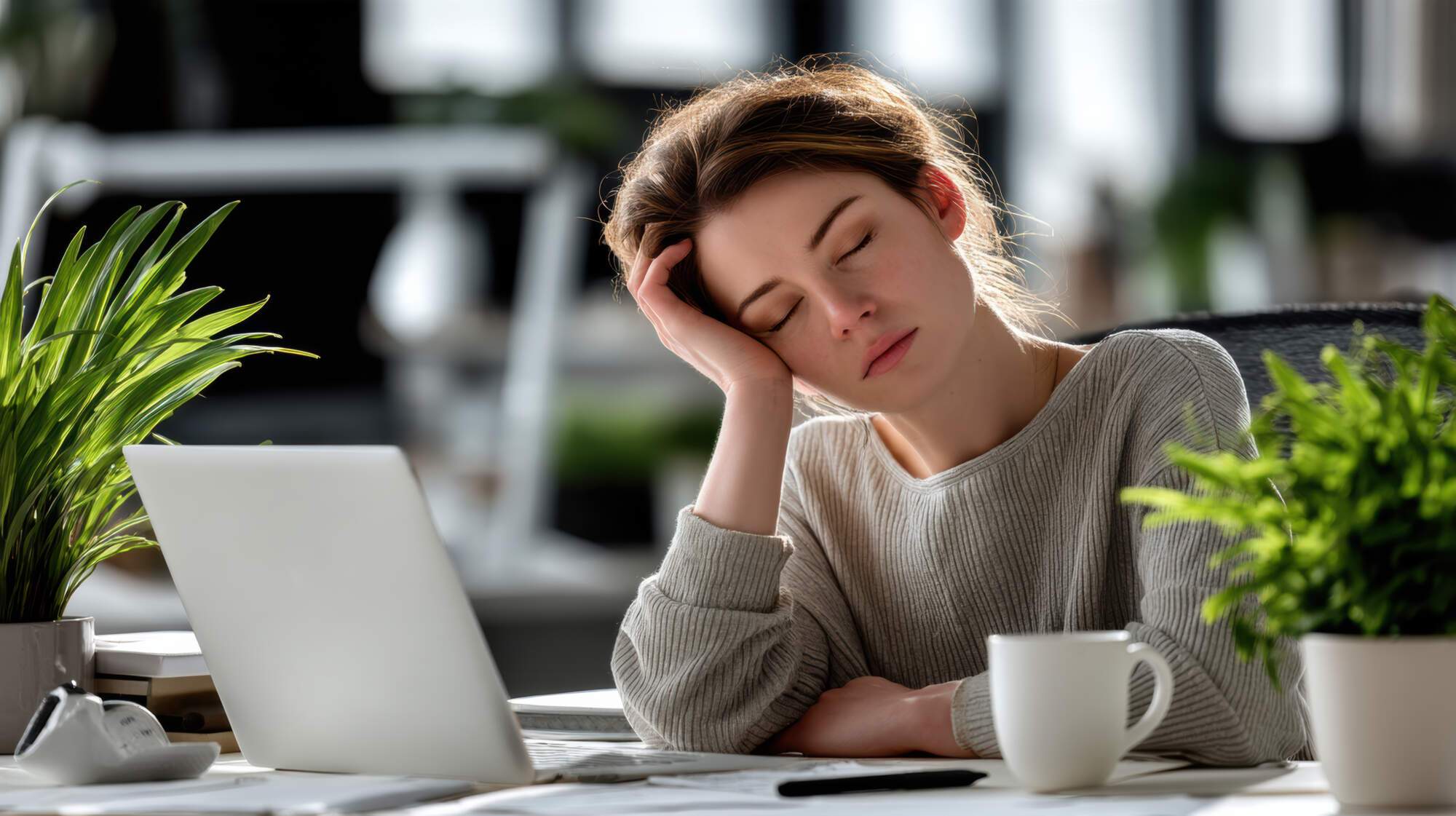 Woman in beige sweater leaning head on hand at desk with laptop, looking exhausted and fatigued